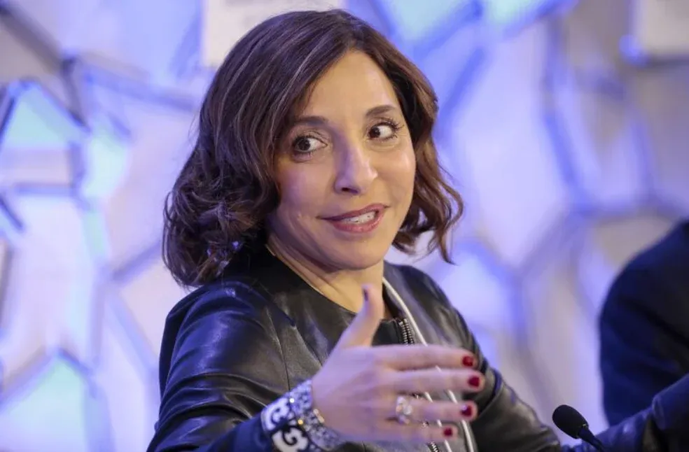 A woman with shoulder-length brown hair and a leather jacket speaks while gesturing with her hands.