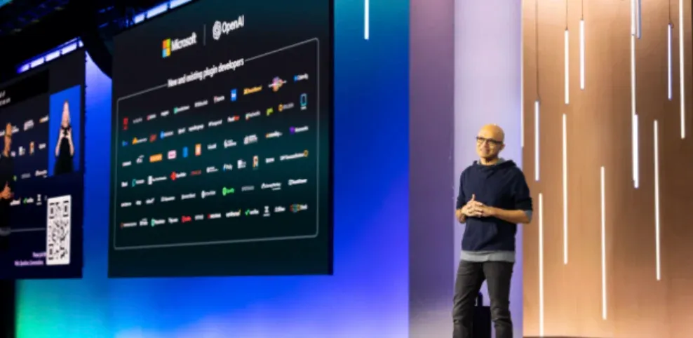 Satya Nadella standing on stage at a Microsoft event with a large screen behind him displaying logos of various plugin developers for Microsoft and OpenAI.